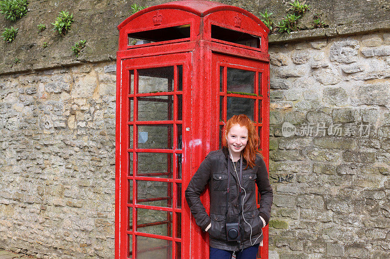 这是一个14 / 15岁的红发少女站在英国街道上一个废弃的红色电话亭旁的图片，她穿着绿色蜡制夹克，蓝色牛仔牛仔裤，格子帆布鞋，脖子上挂着一个相机盒，用耳塞听着音乐